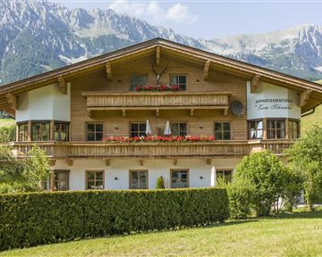 Ein schönes Holzhaus mit Balkonen und blühenden Blumenkästen. Die Umgebung ist von grünen Wiesen und Bergen umgeben.