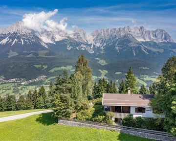 Ein gemütliches Haus in einer idyllischen Landschaft. Im Hintergrund erheben sich majestätische Berge unter einem klaren Himmel.