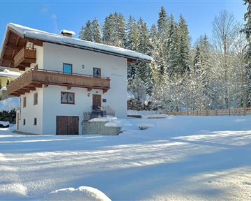 Ein charmantes Haus im Schnee, umgeben von hohen Bäumen. Der Himmel ist klar und blau, was eine winterliche Atmosphäre schafft.