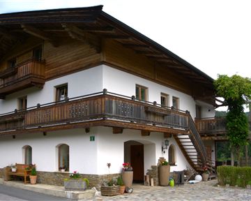A traditional house in Alpine style with a wooden balcony. In the background, green mountains and a clear sky can be seen.