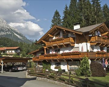 Ein traditionelles Chalet umgeben von Bäumen und Bergen. Die Balkone sind mit Blumen geschmückt, und die Umgebung ist ruhig und malerisch.