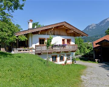 Ein hübsches Haus im alpinen Stil, umgeben von grünem Gras und Bäumen. Im Hintergrund sind Berge sichtbar und der Himmel ist blau.