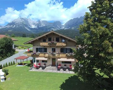 A cozy house in the mountains with blooming balconies. In the background, majestic mountains and a clear blue sky can be seen.