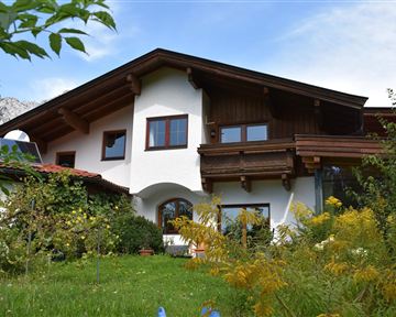 Ein charmantes Haus mit Holzverkleidung und großen Fenstern in einem grünen Garten. Im Hintergrund sind sanfte Hügel sichtbar.
