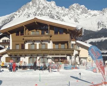 A cozy chalet in the mountains, surrounded by snow. In the foreground are people skiing and relaxing.