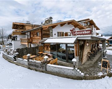 Ein gemütliches Holzhaus im Schnee mit der Beschilderung "Hexenbar". Es gibt Außenplätze zum Sitzen und eine einladende Atmosphäre.