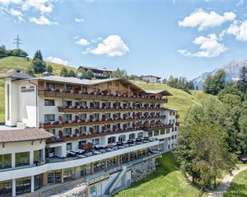 Ein modernes Hotel mit mehreren Balkonen, umgeben von grünen Hügeln und Bergen. Der Himmel ist klar und sonnig.