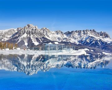 Ein atemberaubender Blick auf schneebedeckte Berge mit einem glitzernden Schwimmbad im Vordergrund. Der klare blaue Himmel spiegelt sich im Wasser wider.