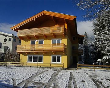 A yellow house with wooden balconies stands on a snow-covered area. In the background, trees and blue sky are visible.