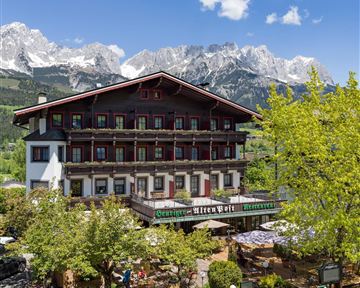 A charming hotel with traditional architecture, surrounded by green trees. Majestic mountains rise in the background beneath a clear sky.