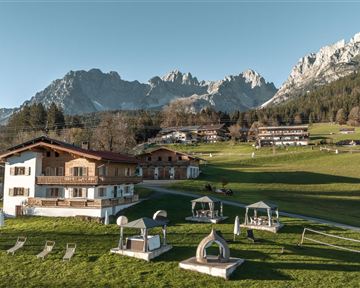 Ein malerisches Alpenresort mit traditionellen Holzgebäuden und einer grünen Wiese. Im Hintergrund erheben sich beeindruckende Berge unter klarem Himmel.