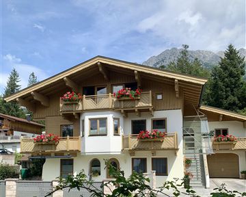 Ein schönes Chalet mit Balkonen, geschmückt mit blühenden Pflanzen. Im Hintergrund sind Berge und ein klarer Himmel zu sehen.