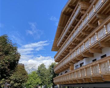 Ein modernes Gebäude mit Holzbalustraden und einem klaren blauen Himmel. Die Straße führt durch eine malerische Landschaft mit Bäumen und Gästen.