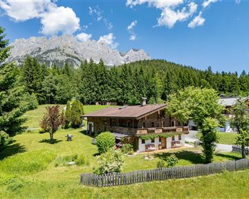 A picturesque alpine landscape with a wooden house and lush greenery. In the background, impressive mountains rise under a blue sky.