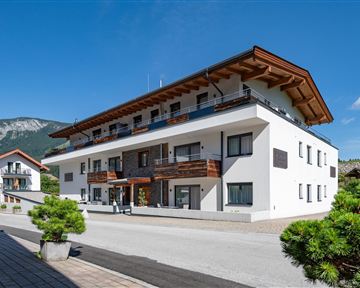 A modern building with several apartments, surrounded by trees and a clear blue sky. In the background, gentle mountains can be seen.