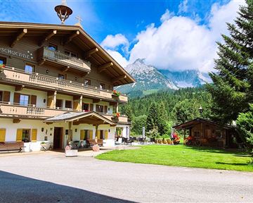 A traditional alpine house with several balconies and beautiful flowers. In the background, green meadows and mountains can be seen.