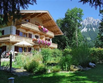 A beautiful wooden house with balconies, adorned with flowers. In the background, mountains covered with trees can be seen.