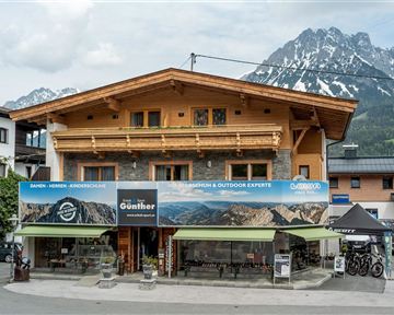 A cozy wooden building with a large advertising sign. In the background, impressive mountains and a partly cloudy sky can be seen.