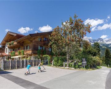 A charming wooden building is located in a picturesque landscape. Two children are riding their scooters along a road in front of the building.