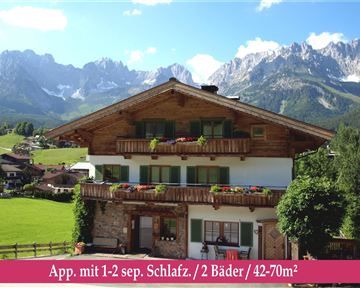 Ein schönes Chalet in den Bergen mit blühenden Balkonen und beeindruckender Aussicht. Im Vordergrund ist ein grünes Feld zu sehen, das die ländliche Idylle unterstreicht.