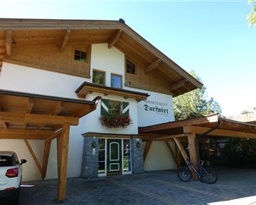 Ein gemütliches Apartmenthaus mit Holzverkleidung und blühenden Pflanzen. Im Vordergrund stehen ein Auto und ein Fahrrad.
