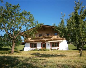 Ein traditionelles, zweigeschossiges Haus im bayerischen Stil mit Holzverandabalkon. Umgeben von grünen Wiesen und Bäumen unter klarem Himmel.