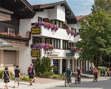 Eine malerische Straße mit traditionellen Gebäuden und bunten Blumenkästen. Menschen gehen und fahren mit dem Fahrrad vorbei, während Bäume Schatten spenden.