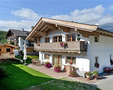 Ein gemütliches alpenländisches Haus mit einem Balkon und Blumenkübel. Der Garten ist grün und gepflegt, umgeben von Bergen und einem klaren Himmel.
