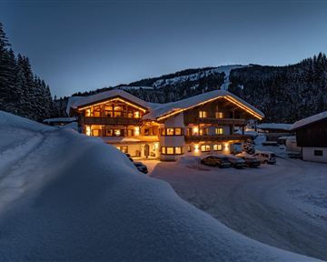 Ein gemütliches Chalet im Schnee, beleuchtet von warmem Licht. Die Umgebung ist von Bäumen und Bergen umgeben.