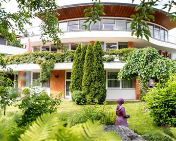 Ein modernes Gebäude mit einem runden Balkon, umgeben von üppiger Vegetation. Im Vordergrund steht eine lila Statue in einem gepflegten Garten.