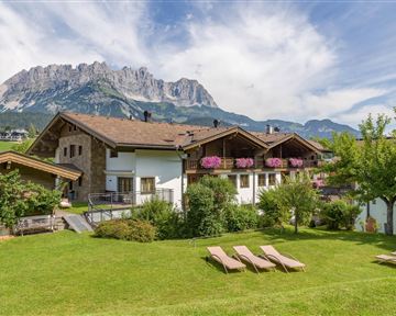 Ein ruhiges Chalet im Grünen mit blühenden Blumen. Im Hintergrund erheben sich majestätisch die Berge unter einem klaren Himmel.