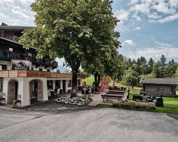 Ein gemütliches Gasthaus in einer grünen Umgebung. Vor dem Gebäude befinden sich Sitzbereiche und ein großer Baum.