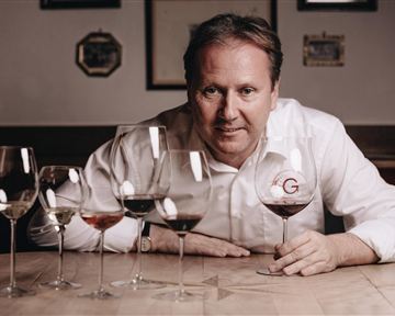 Ein Mann sitzt an einem Tisch mit mehreren Weingläsern. Er hält ein Glas Wein in der Hand und lächelt.