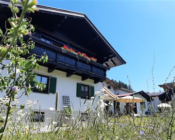 Ein traditionelles Haus mit einem Balkon voller Blumen und einem großen Garten. Die Sonne scheint an einem klaren blauen Himmel.