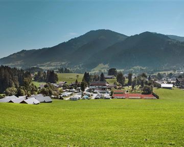 Eine malerische Landschaft mit sanften Hügeln und einem kleinen Dorf. Im Hintergrund sind Berge und ein blauer Himmel zu sehen.