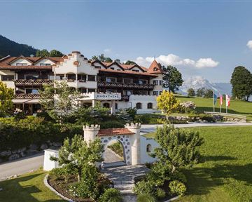 Ein charmantes Hotel mit traditioneller Architektur, umgeben von grünen Wiesen und Bäumen. Im Hintergrund sind Berge und ein klarer Himmel sichtbar.