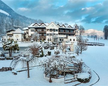 Ein idyllisches Hotel im Schnee, umgeben von verschneiten Bäumen und Bergen. Der Himmel ist leicht bewölkt und verleiht der Szene eine ruhige Atmosphäre.