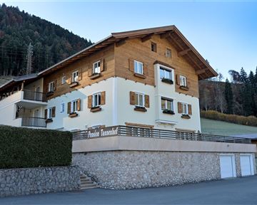 Ein charmantes, zweigeschossiges Haus mit Holzverkleidung und Fensterläden. Es liegt in einer malerischen Landschaft umgeben von Wäldern.