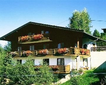 Ein gemütliches Holzhaus mit Balkonen und bunten Blumen. Es liegt in einer grünen Umgebung und hat einen klaren blauen Himmel im Hintergrund.