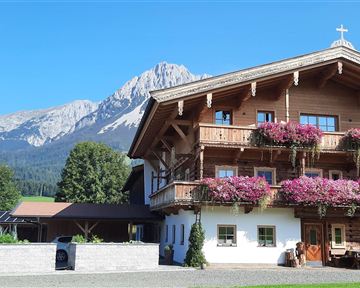 A cozy wooden house with blooming balcony flowers in front of an impressive mountain landscape. The sky is clear and the nature around the house is green and inviting.