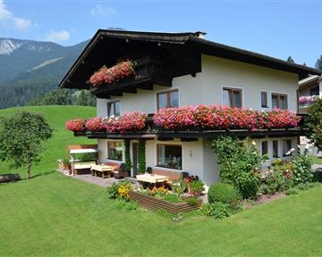 Ein charmantes Haus mit blühenden Balkonblumen in einer grünen Umgebung. Im Hintergrund sind sanfte Hügel und Berge sichtbar.