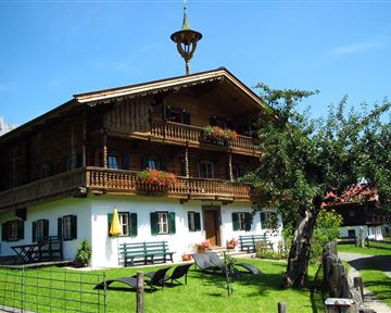 Ein traditionelles alpenländisches Holzhaus mit einem Balkon und bunten Blumen. Im Vordergrund gibt es eine grüne Wiese und einige Möbel zum Entspannen.