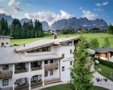 Ein gemütliches Hotel in den Bergen, umgeben von grünen Wiesen und hohen Bäumen. Im Hintergrund sind beeindruckende Berge zu sehen.