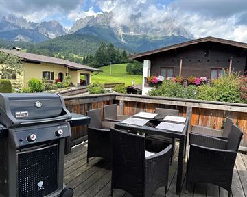 A terrace with a grill and a table, surrounded by comfortable chairs. In the background, impressive mountains can be seen under a partially cloudy sky.