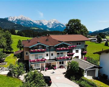 Ein malerisches Haus in der Natur mit Blumenkästen an den Balkonen. Im Hintergrund sind die majestätischen Berge und ein blauer Himmel zu sehen.
