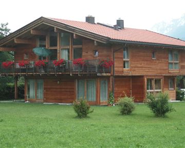 Ein gemütliches Holzhaus mit einem roten Dach und Balkonen, die mit Blumen geschmückt sind. Um das Haus herum befindet sich ein grüner Rasen und Berge im Hintergrund.