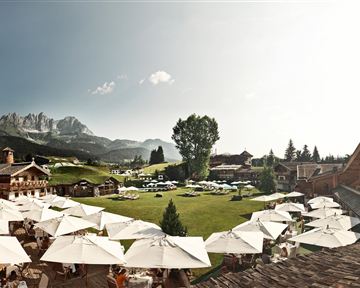 Ein schönes Hotel mit einem großen Garten und Sonnenschirmen. Im Hintergrund sind Berge und ein klarer Himmel zu sehen.