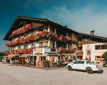 A charming alpine hotel with blooming flower balconies. In front of the building, there is a white car parked in the parking lot.