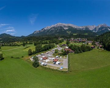 Eine weite, grüne Landschaft mit Bergen im Hintergrund. In der Mitte sind Wohnmobile und ein kleiner Ort sichtbar.