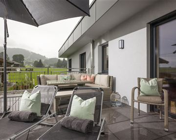 Eine moderne Terrasse mit bequemen Liegen und einem Sofa. Im Hintergrund sind grüne Wiesen und bewölkter Himmel zu sehen.